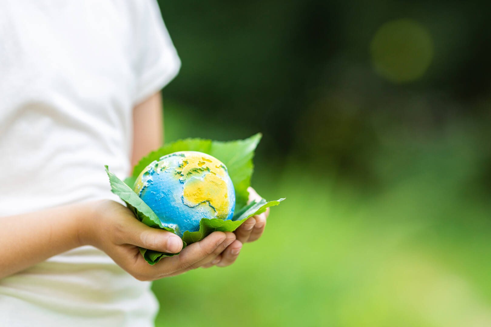 Kid holding small planet in hands against spring or summer green background. Ecology, environment and Earth day concept.